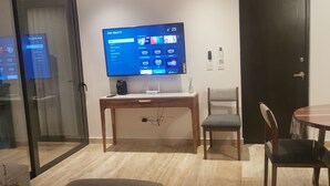 A modern living room with a flat-screen TV mounted on the wall, a wooden desk, a chair, and a dining table with chairs. -  Playa Condo with  Rooftop pool & ocean view - walking  to famous 5ta & Beach. (Playa del Carmen)