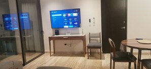 A modern living room with a flat-screen TV mounted on the wall, a wooden desk, a chair, and a dining table with chairs. -  Playa Condo with  Rooftop pool & ocean view - walking  to famous 5ta & Beach. (Playa del Carmen)