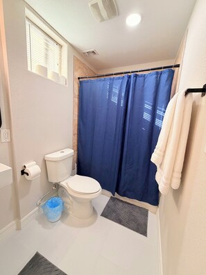 A bathroom with a toilet, blue shower curtain, white towel rack, and a blue trash bin. - Nice 2-bedroom house with AC in welcoming Houston neighborhood (Houston)