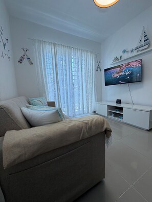 A modern living room with a sofa, a television mounted on the wall, and a window with sheer curtains. - The Best Location in Bayahibe (Bayahíbe)