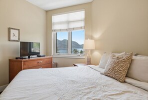 Ein Schlafzimmer mit einem Bett, einem Fernseher auf einer Holzkommode, einer Lampe und einem Fenster mit Blick auf Berge.