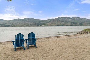 Zwei Adirondack-Stühle an einem sandigen Strand, im Hintergrund ein See und Berge.