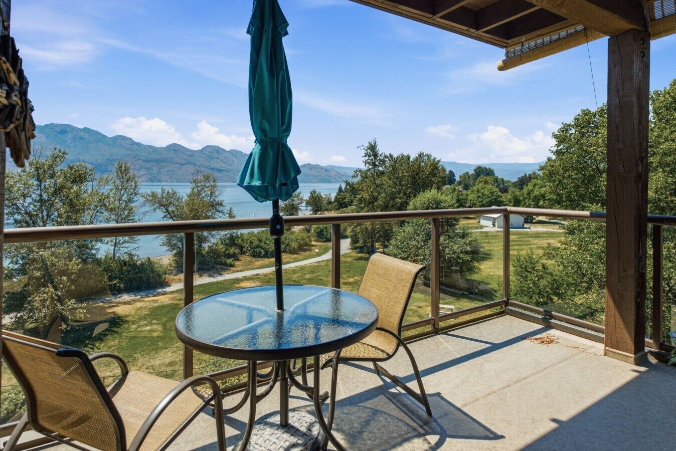 A balcony with a glass table and chairs, a green umbrella, and a view of a lake and mountains.