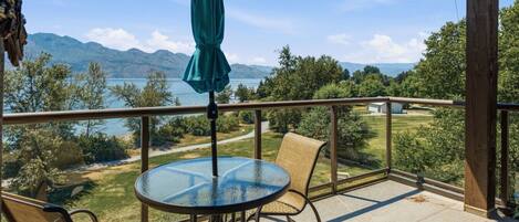A balcony with a glass table and chairs, a green umbrella, and a view of a lake and mountains.