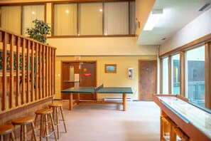 A room with a ping pong table, wooden stools, a potted plant, and a view through large windows. - Ski-In/Ski-Out Studio | Mountain Views (Big White Mountain)