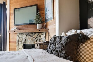 A bedroom with a stone fireplace, a television mounted above it, and a bed with a checkered pillow. - Ski-In/Ski-Out Studio | Mountain Views (Big White Mountain)