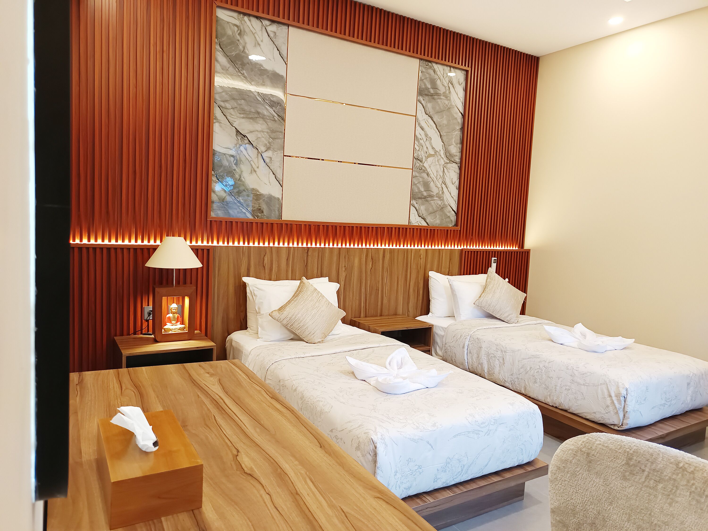 A hotel room with two beds, a wooden nightstand with a lamp, a wooden table, and a large marble artwork on the wall.