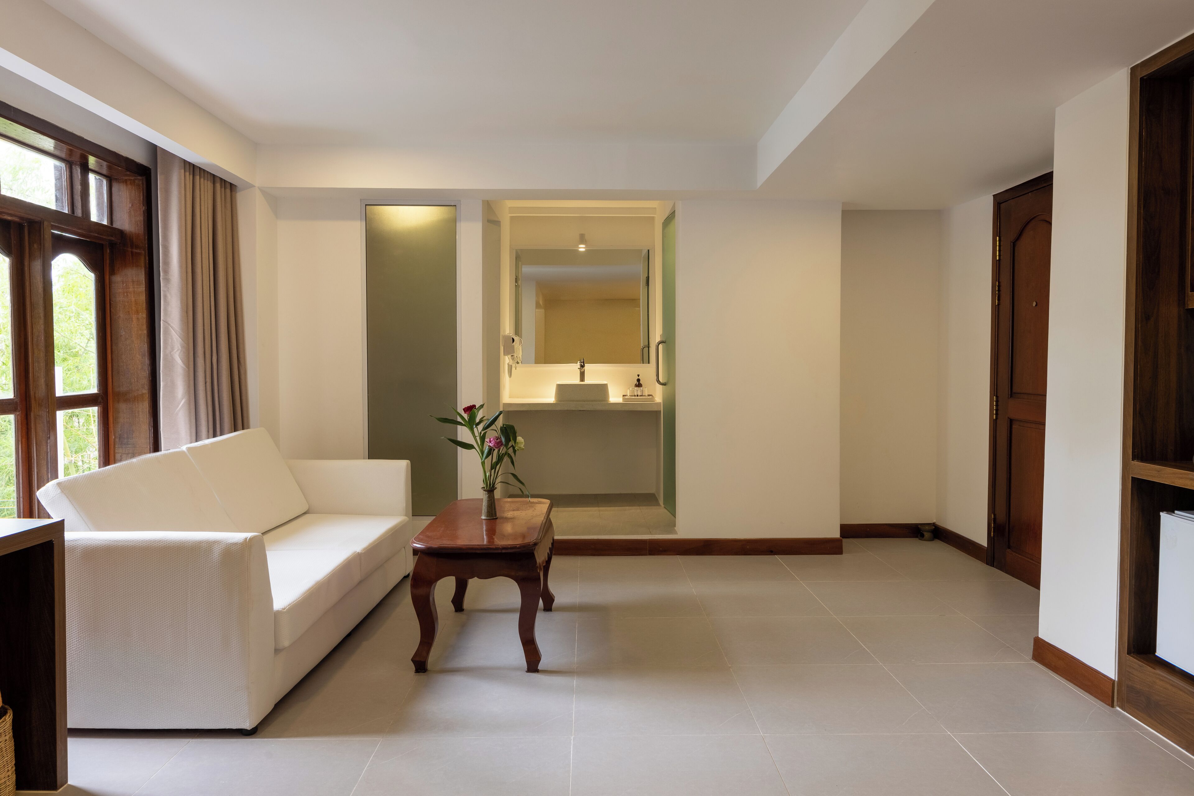 Una sala de estar moderna con un sofá blanco, una mesa de centro de madera y vista al baño por una puerta de vidrio.