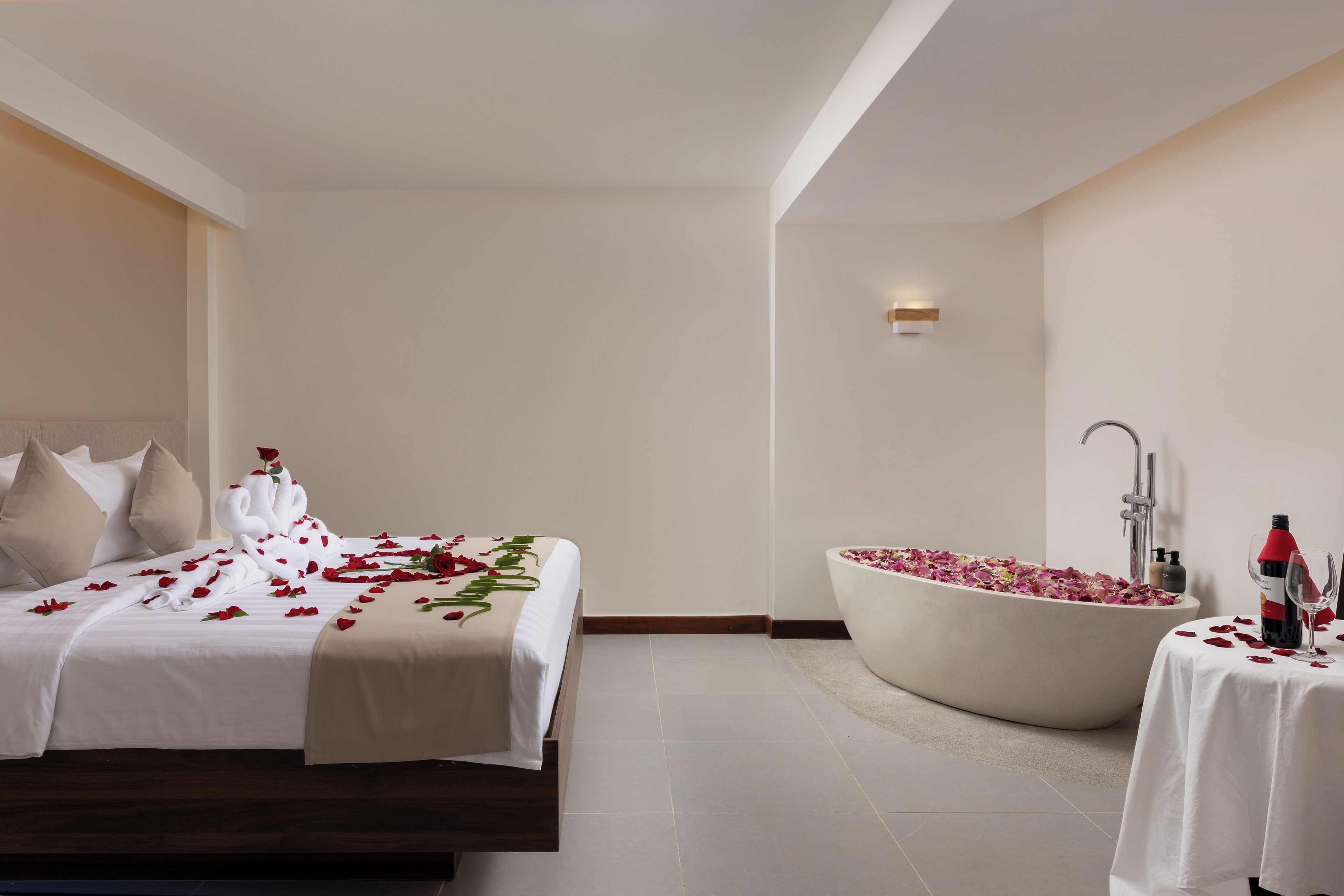 Habitación de hotel con una cama grande, una bañera llena de pétalos de rosa y una mesita con una botella y un vaso.