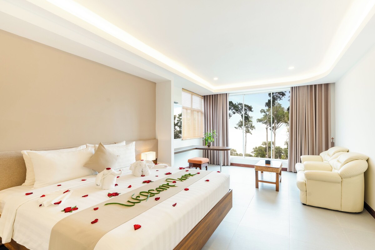 A hotel room with a large bed, a sofa, a small table, and a view of trees through the window.