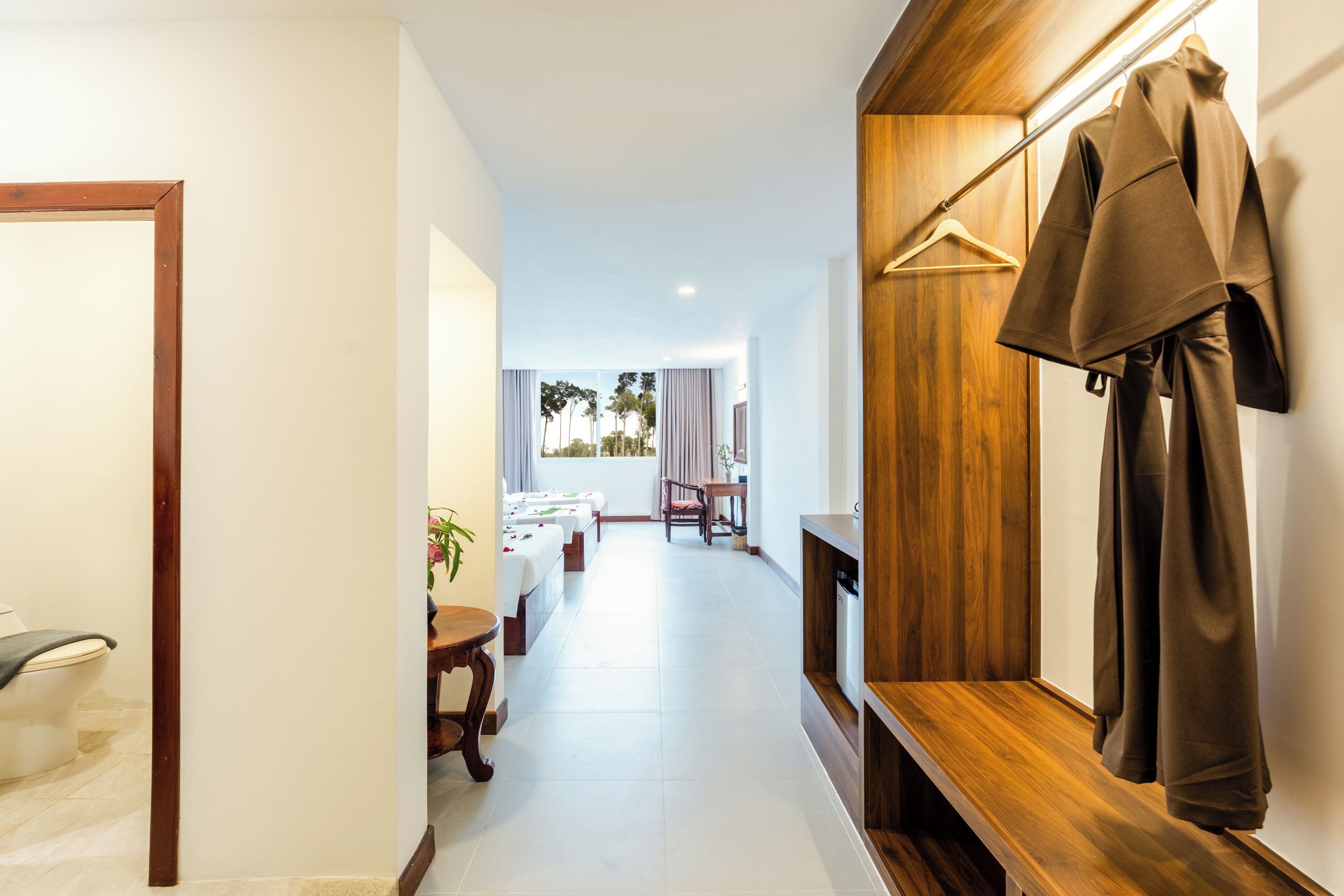 A hotel hallway with a wooden bench, a coat rack, and a view of a dining area with tables and chairs.