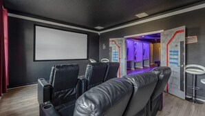 Una sala cinematografica con poltrone in pelle nera reclinabili, uno schermo grande e vuoto e un corridoio illuminato di viola.