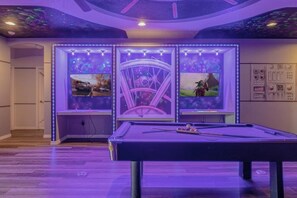 Une table de billard dotée d’un éclairage à LED, un plafond orné d’une fresque sur le thème de l’espace, et deux écrans encadrés diffusant des jeux vidéo.