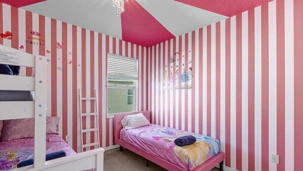 Camera da letto per bambini con pareti a righe rosa e bianche, un letto a castello, una finestra con persiane e un letto con coperta colorata.