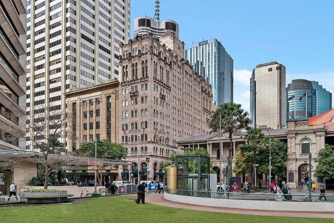 Une place de ville bordée de bâtiments historiques, d’un immeuble moderne en verre et d’une place centrale dotée d’un espace aménagé avec des sièges.