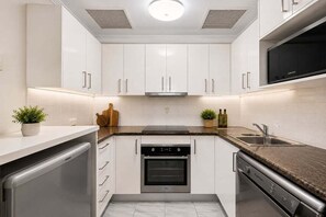 A compact kitchen with white cabinetry, a built-in oven, a dishwasher, and a microwave. - Modern CBD Retreat | Walk to Shops & Dining (Brisbane)
