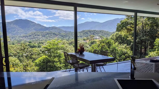 A dining area with a table and chairs, a vase with flowers, and a view of a mountainous landscape through large windows. - Luxury 3 bedroom Villa with Private Pool Views (Edge Hill)