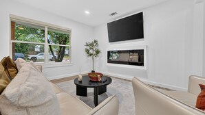 Una sala de estar moderna con un sofá, una mesa de centro, un televisor montado en la pared y un ventanal con vistas al exterior.