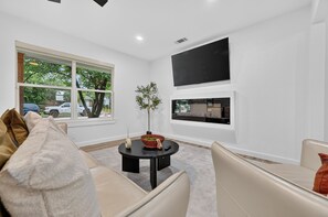 A modern living room with a sofa, a coffee table, a TV mounted on the wall, and a large window providing a view of the outside. - Executive 3BR Home, King Suite, Near Hospitals & Historic Downtown Mansfield (Mansfield)