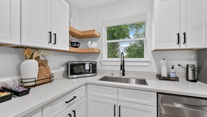 Cocina moderna con armarios blancos, un lavamanos de acero inoxidable, microondas y ventana con vistas a árboles.