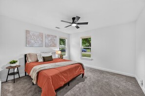 Ein Schlafzimmer mit einem Bett, einem Deckenventilator, zwei Fenstern und Kunstwerken an den Wänden.
