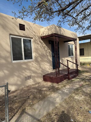 A single-story building with a beige stucco exterior, a front porch with a wooden railing, and a small set of steps leading to the entrance. - Charming Retreat in ABQ's North Valley-close to Old Town and Balloon Fiesta Park (Albuquerque)