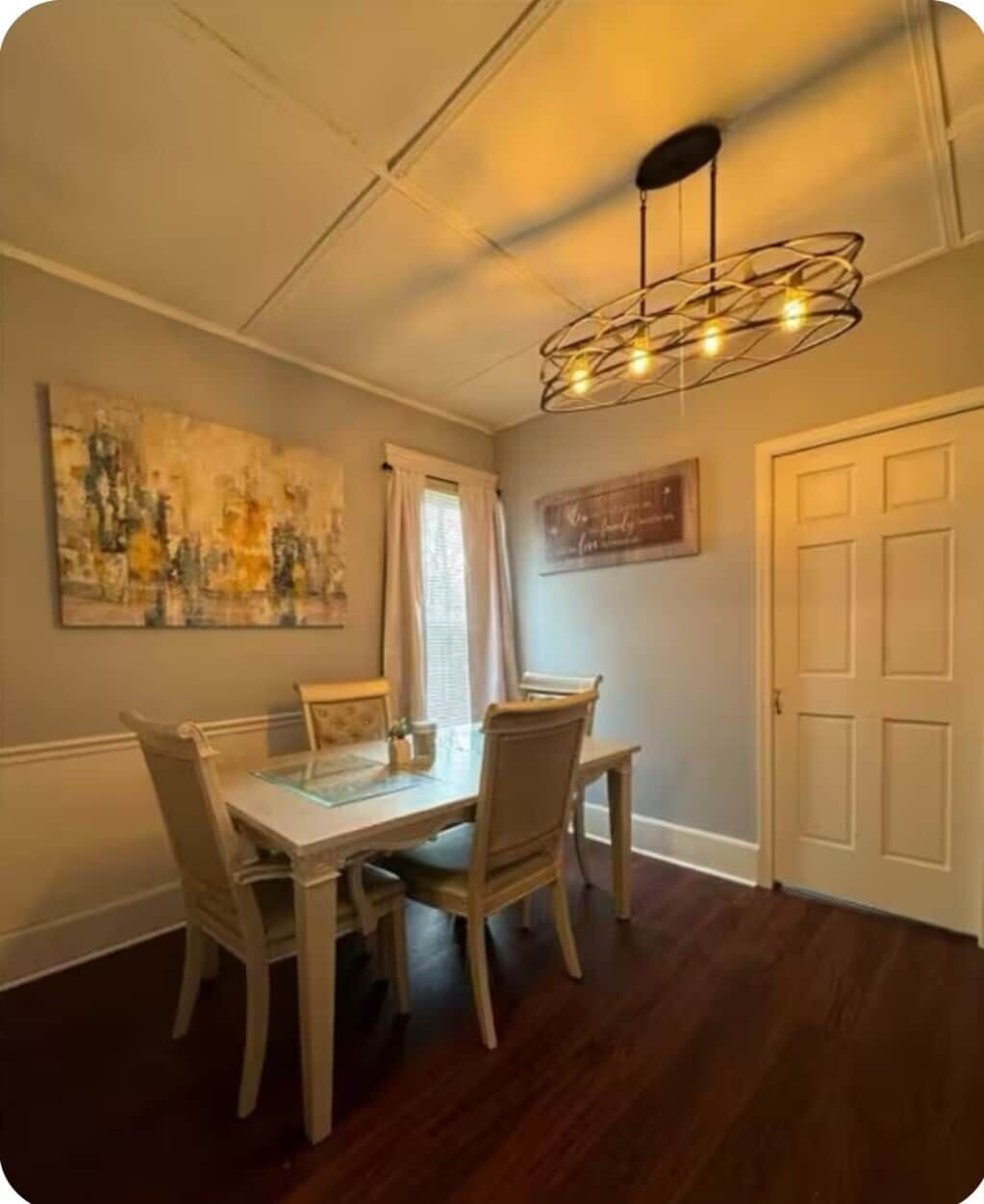 Un coin repas comprenant une table à plateau de verre, des chaises en bois, un lustre moderne, un tableau accroché au mur et une porte fermée.
