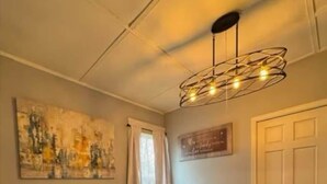 Un coin repas comprenant une table à plateau de verre, des chaises en bois, un lustre moderne, un tableau accroché au mur et une porte fermée.