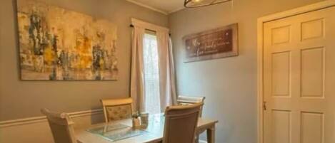 Un coin repas comprenant une table à plateau de verre, des chaises en bois, un lustre moderne, un tableau accroché au mur et une porte fermée.