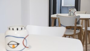 Zona pranzo con tavolo e sedie bianche, una tazza decorativa in ceramica e una finestra con vista all'esterno.