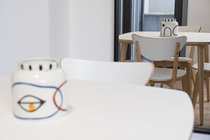 Ein Essbereich mit weißem Tisch und Stühlen, einer dekorativen Keramiktasse und einem Fenster mit Blick nach draußen.