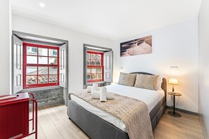 A modern bedroom with a bed, bedside lamp, and a view through a window with red frames. - GuestReady - Cosy place in Braga's historic heart (Braga)