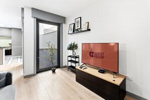 Une chambre d’hôtel moderne dotée d’une porte coulissante donnant sur un balcon, d’un téléviseur à écran plat posé sur une console en bois et d’une petite plante sur une étagère.
