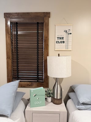 A bedroom with a wooden window blind, a framed welcome sign, a bedside lamp, a golf book, and a small plant on the nightstand.