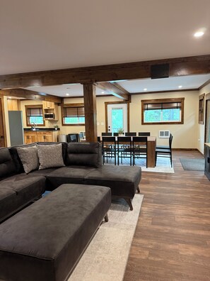 A spacious living area with a sectional sofa, a dining table with chairs, a kitchen in the background, and a large window with a view of trees.