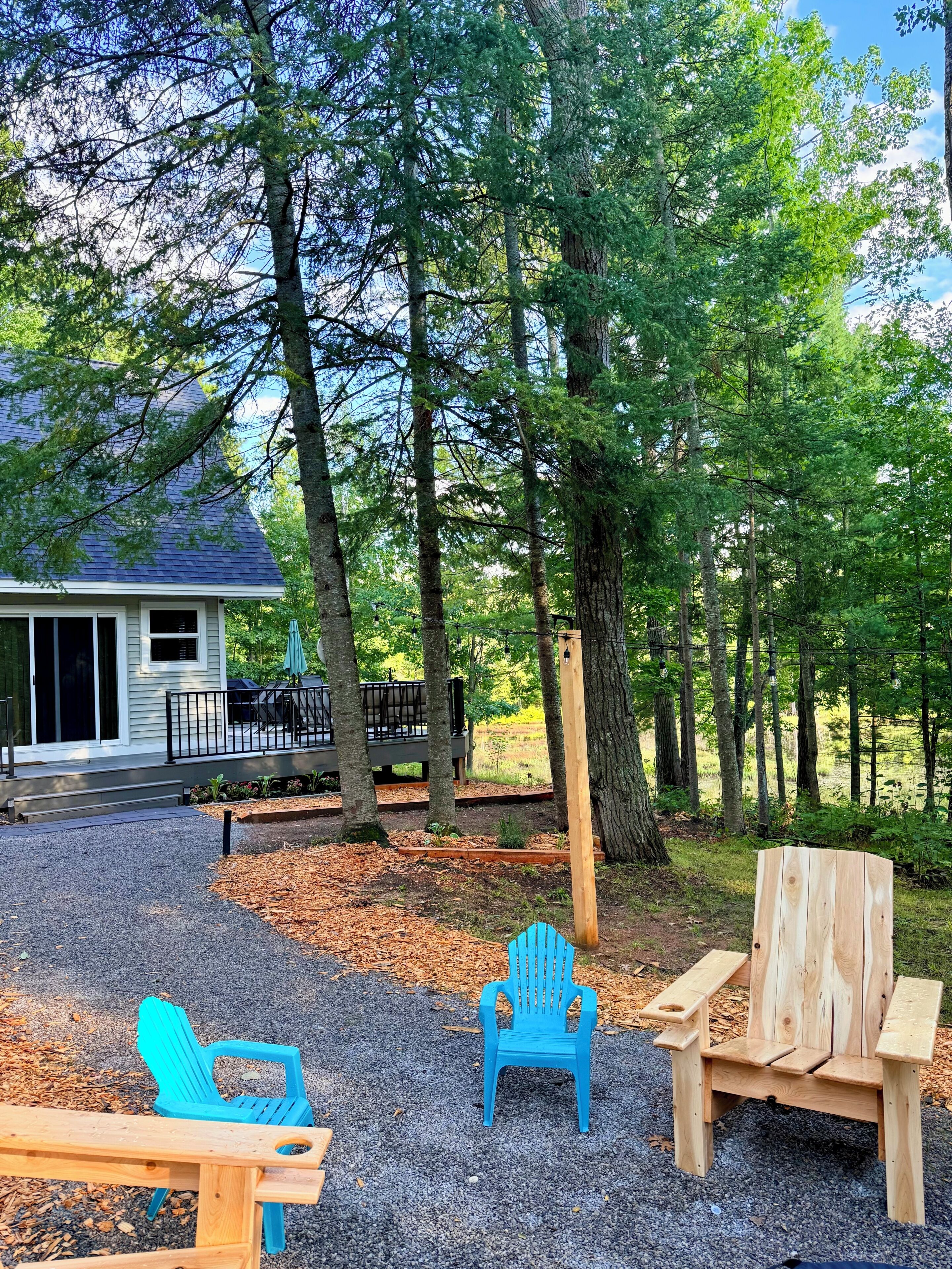 Una terraza de madera con sillas Adirondack y una mesa de picnic, rodeada de árboles y una casa con terraza.