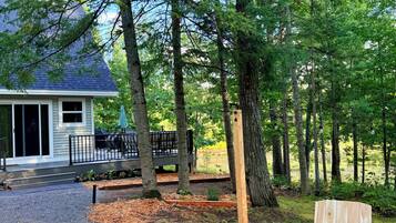 Una terraza de madera con sillas Adirondack y una mesa de picnic, rodeada de árboles y una casa con terraza.