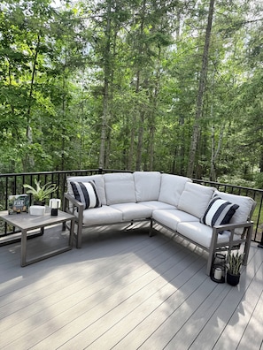 A white outdoor sectional sofa with metal legs on a wooden deck, surrounded by a metal railing, with a book and a plant on the table beside it.