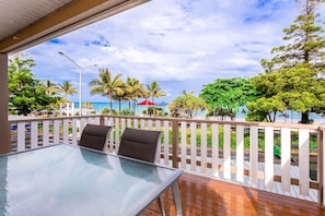 A glass-top table with two chairs, a wooden deck, a white picket fence, and a view of the ocean and palm trees. - Wake to Sunrises on Yeppoon’s Beachfront (Yeppoon)