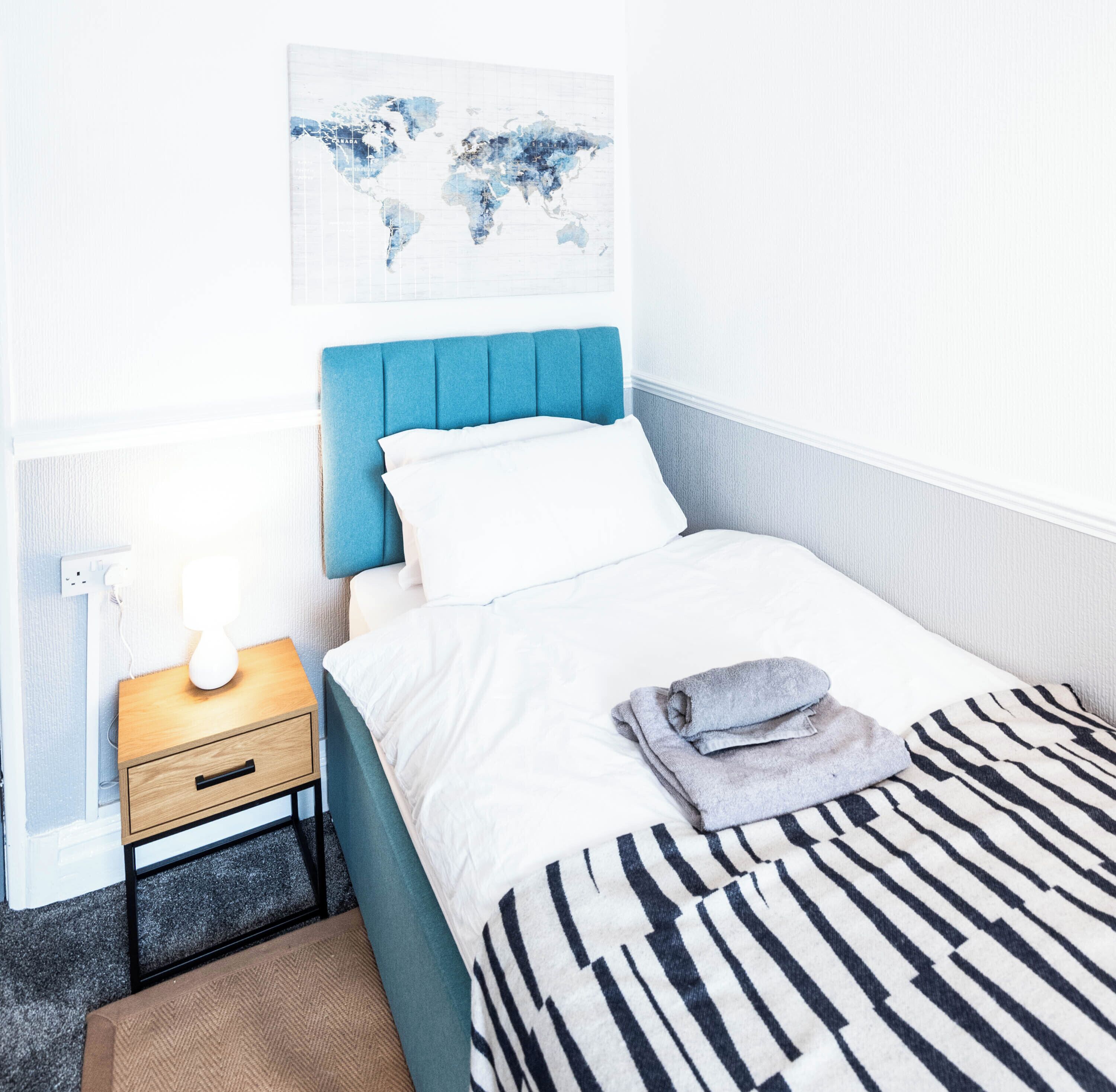 A neatly made bed with a striped blanket, a world map artwork on the wall, a bedside table with a lamp, and a small drawer.