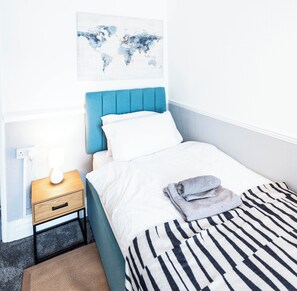 Een net opgemaakt bed met een gestreept kleed, een wereldkaart aan de muur, een nachtkastje met een lamp en een klein ladeblok.