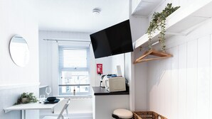 Ein kompakter, moderner Raum mit Flachbildfernseher, einem kleinen Tisch mit Schüssel und Weinglas, einem Stuhl, einer Heizung, einer weißen Wand und einem Fenster mit Jalousien.