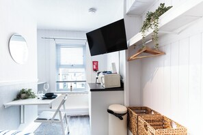 Ein kompakter, moderner Raum mit Flachbildfernseher, einem kleinen Tisch mit Schüssel und Weinglas, einem Stuhl, einer Heizung, einer weißen Wand und einem Fenster mit Jalousien.