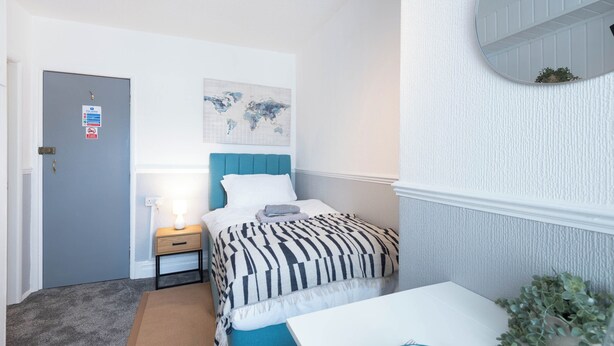 Ein Schlafzimmer mit einem Bett, einem Nachttisch, einer Lampe, einer Weltkarte an der Wand, einer Tür mit Schildern und einer kleinen Pflanze.