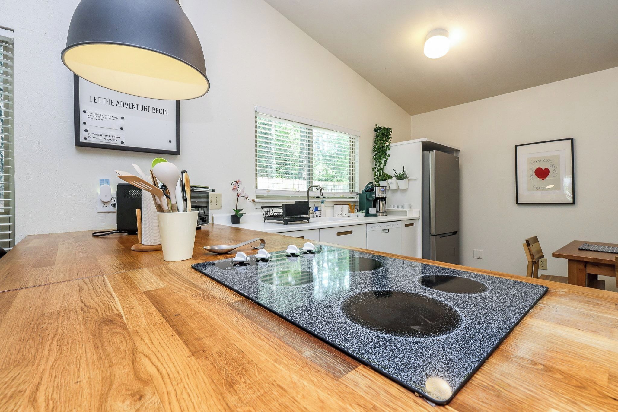 A modern kitchen with a central island, a black pendant light, a framed inspirational quote, and a refrigerator.