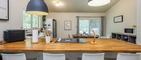 A modern kitchen with a wooden island, white chairs, pendant lights, and a dining area with a view of trees.