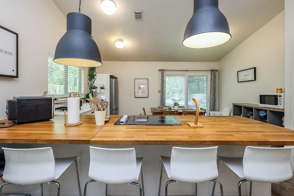 A modern kitchen with a wooden island, white chairs, pendant lights, and a dining area with a view of trees.