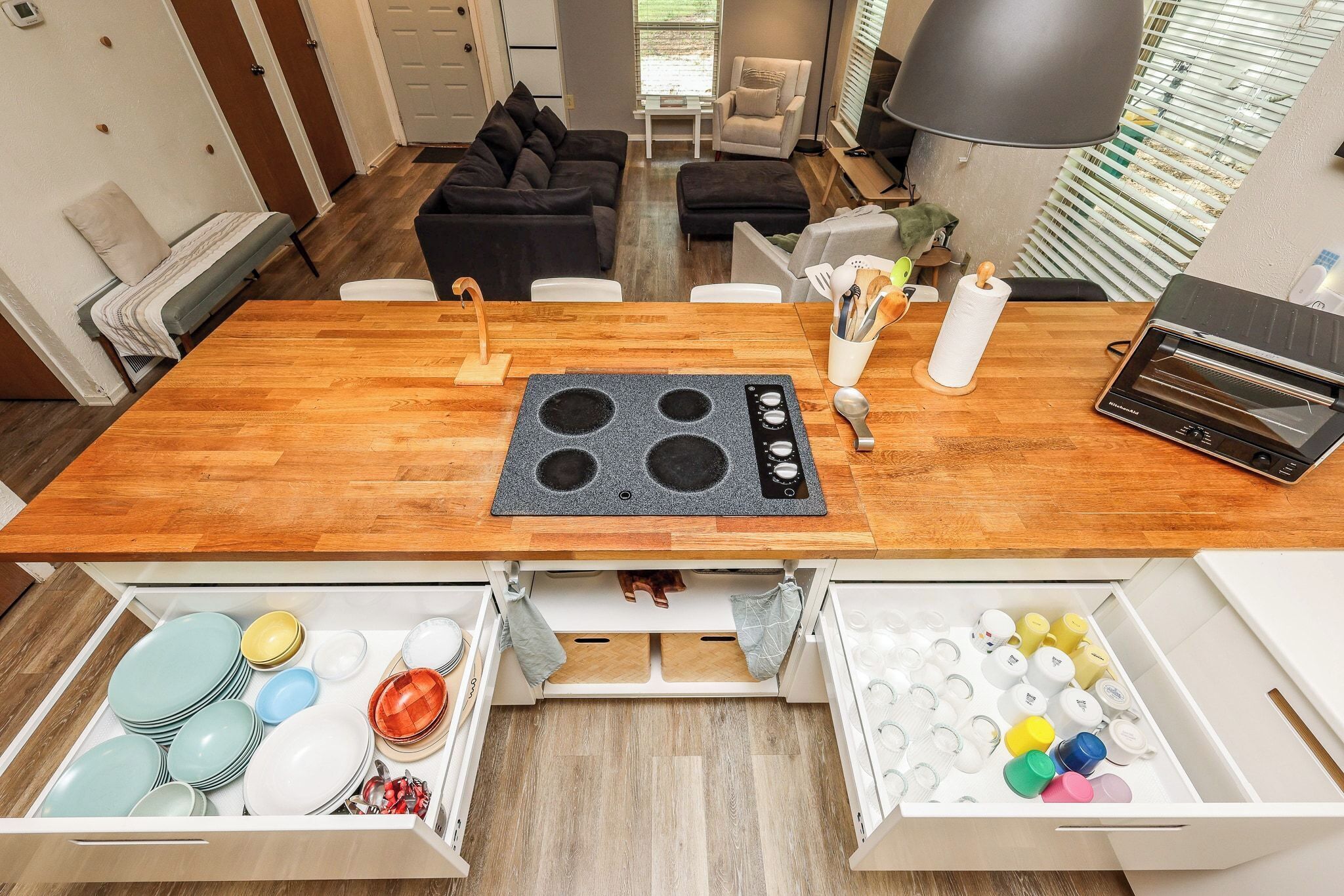 A kitchen with a built-in stove, wooden table, and two open storage drawers with dishes and utensils.