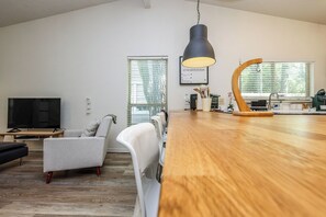 A modern living room with a flat-screen TV, a dining area with a wooden table, and a kitchen in the background.