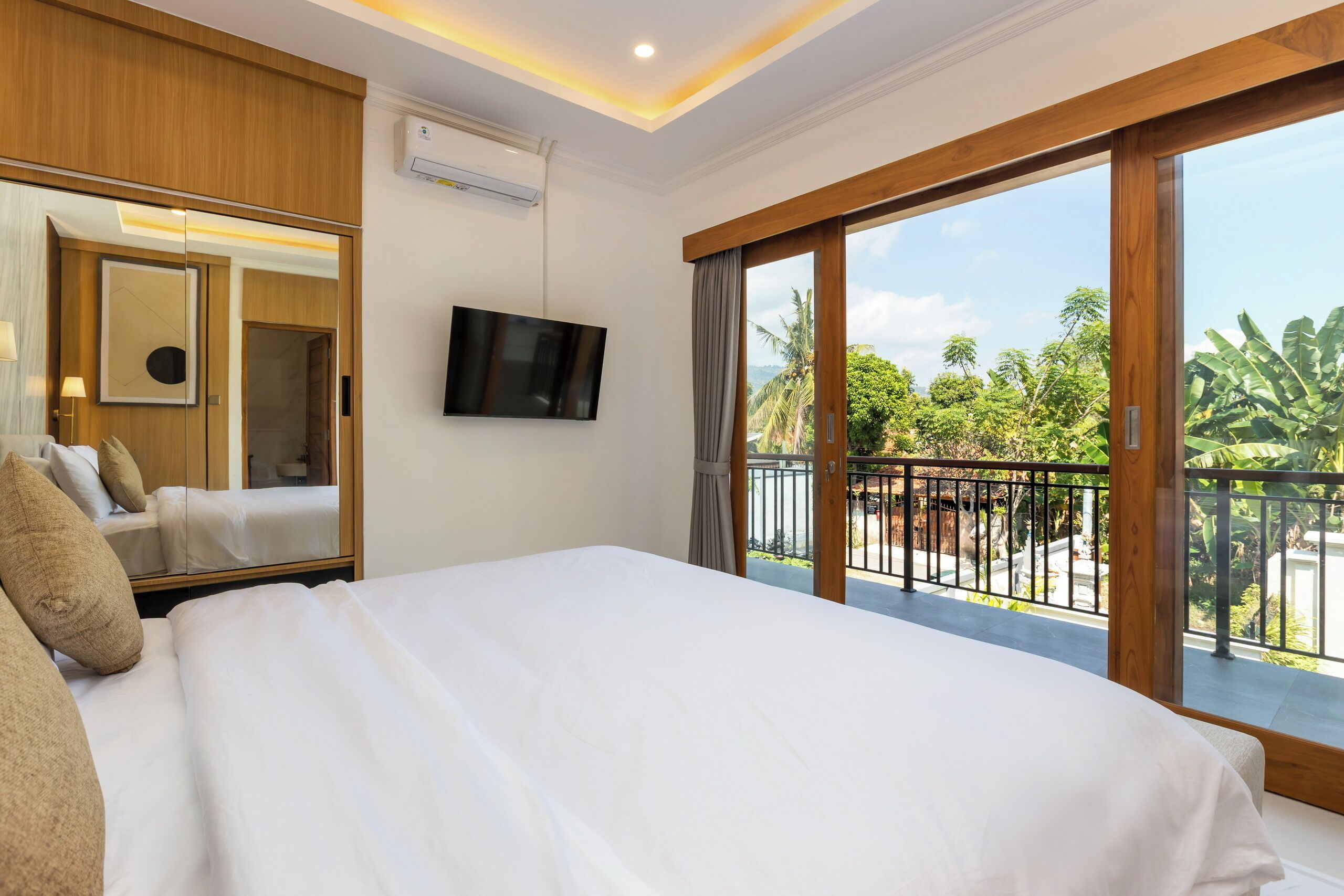Habitación de hotel con una cama grande, televisión, espejo y balcón con vista a la vegetación.